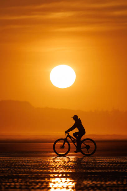 A woman goes for a bike ride at low tide on Long Beach on Vancouver Island near Tofino, British Columbia, Canada, at sunset. She wears casual clothes and a sun hat.