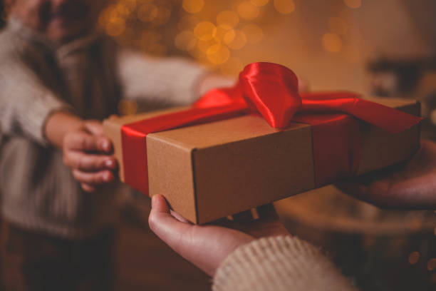 istock Hands exchanging a festive gift box with a red ribbon under a beautifully decorated Christmas tree. Cozy holiday setting with candles, miniature houses, and a warm atmosphere 2194576126