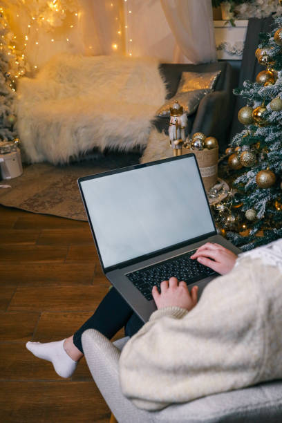 istock Close-up of a laptop with a blank white screen, hands typing on the keyboard, with a blurred background of a lit Christmas tree and festive decorations, creating a cozy holiday atmosphere 2194576129