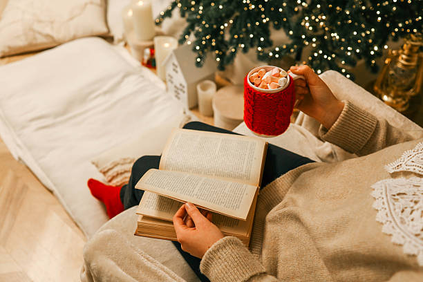 A cozy holiday scene featuring a person reading an open book while holding a festive red mug filled with marshmallows