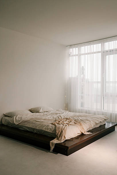 istock Minimalist bedroom with a cozy unmade bed, soft beige tones, and large windows draped with sheer curtains, letting in natural light. Tranquil and serene interior 2194576467