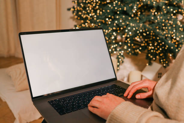 istock Close-up of a laptop with a blank white screen, hands typing on the keyboard, with a blurred background of a lit Christmas tree and festive decorations, creating a cozy holiday atmosphere 2194576479