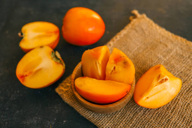 Rustic composition with fresh persimmons in a wooden bowl and on a dark textured background, accented by burlap. Ideal for seasonal, culinary, and rustic lifestyle themes