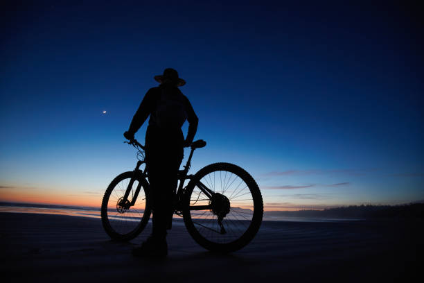 A female cyclist looks at the view of the ocean and the moon on Long Beach near Tofino on Vancouver Island, British Columbia, Canada, at night. She stands next to her mountain bike and wears casual clothing and a sun hat.