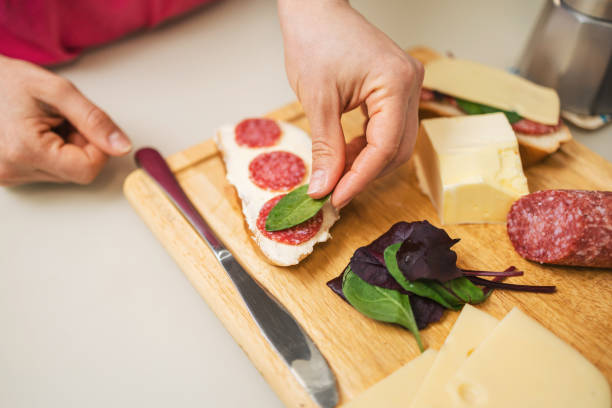 Woman preparing breakfast in a kitchen, making a sandwich with butter, sausage, cheese, and spinach leaves on bread, on a table