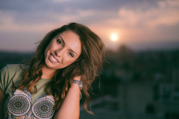 Young woman with casual style smiling warmly at the camera with a background of an urban sunset. The image conveys a sense of relaxation and joy amidst a picturesque outdoor setting