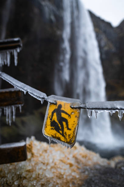 A frozen warning sign hangs from a railing beside a waterfall, indicating slippery terrain. Icicles dangle from the edges, highlighting the cold, icy conditions in the area.