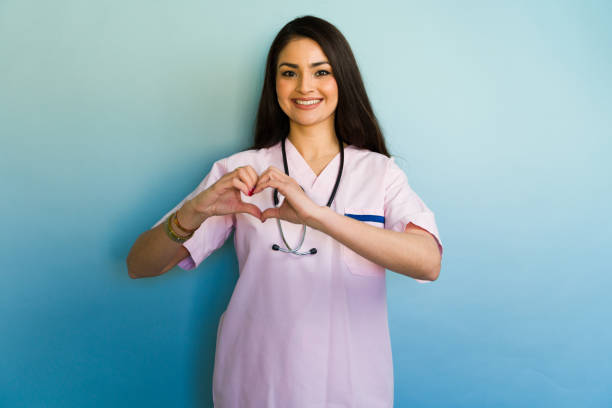 Beautiful young doctor wearing pink scrubs making a heart shape with her hands and smiling at the camera