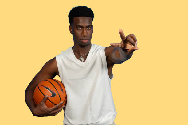 Young man wearing white shirt holding basketball and gesturing with hand on yellow background
