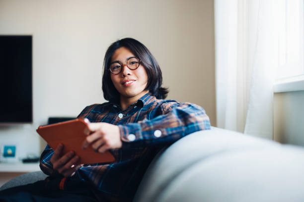 Young person sitting comfortably on a sofa, holding a tablet, while enjoying a casual moment in a well-lit home environment.