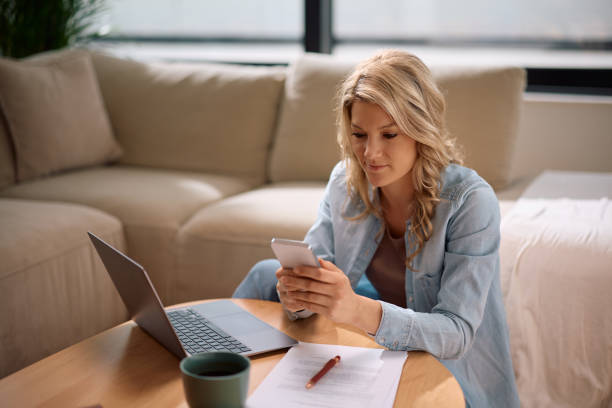 istock Smiling woman texting on cell phone while working on laptop at home. 2194605133