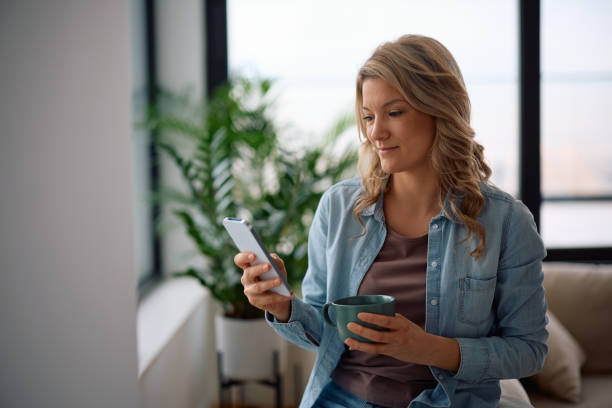 istock Smiling woman using mobile phone while drinking coffee at home. 2194605169