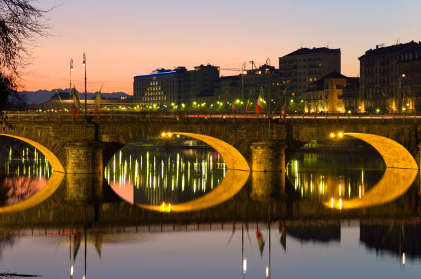 In winter, the night comes early in Turin. Here is the bridge over the Po river, connecting the huge Vittorio Square to the Great Mother church. The calm waters of the river Po reflect the lights of the bridge and the sunset colors. The city its famous for its metaphysical look of lonely squares, equestrian statues and long porticoes, so appreciated by philosopher Nietzsche and painter De Chirico. In the background are the mountains.