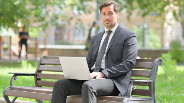 istock Middle Aged Businessman Looking at Camera while Using Laptop Outdoor 2194608784