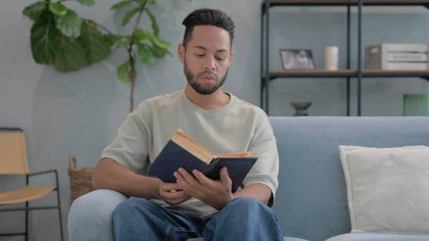 istock Hispanic Man Reading Book while Sitting on Sofa at Home 2194612671