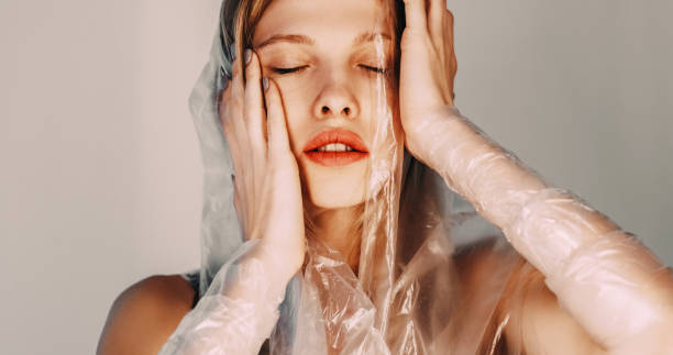 Mental disorder. Woman depression. Tired female wearing transparent polyethylene film touching hair on beige illuminated art background.