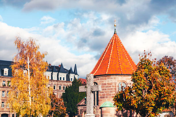 Serene view of Nuremberg Cemetery featuring an old stone cross, autumn trees, and a historic chapel with a red-tiled roof against dramatic sky. Perfect for themes of heritage, history, and tranquility