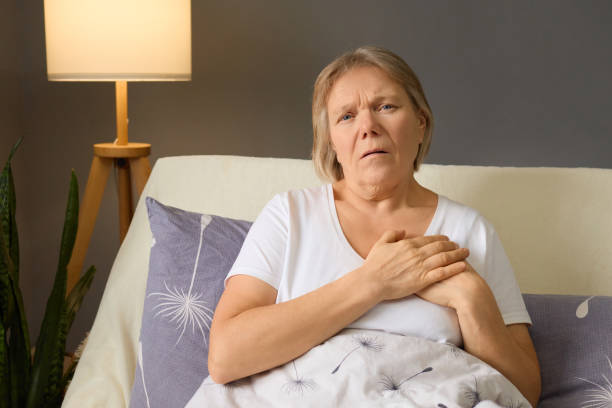istock An elderly woman sits on a bed with a concerned expression, clutching her chest. Her bedroom features soft lighting, plants, and comforting decor, hinting at her discomfort 2194629290