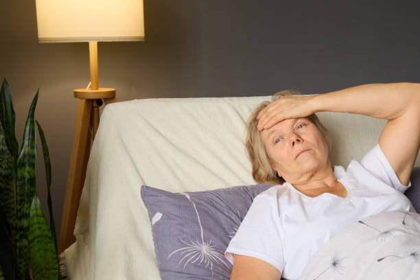 istock A senior woman lies on her bed with a concerned expression, holding her forehead, indicating discomfort. Soft lighting creates a warm, inviting atmosphere 2194629319