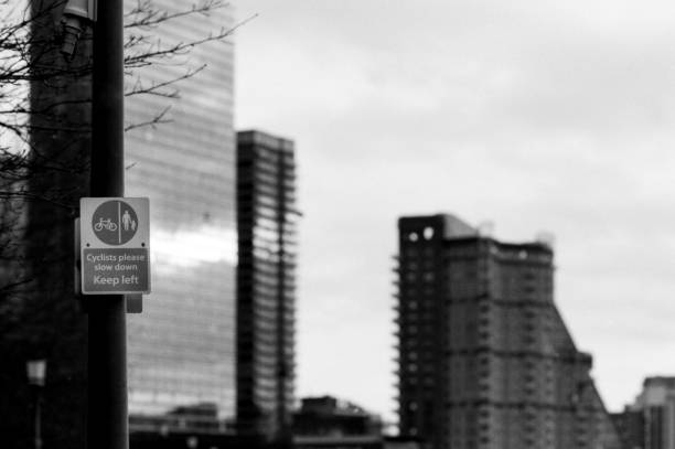 Grainy monochrome analog film capturing a cyclists please slow down, keep left sign in the foreground, with blurred canary wharf skyscrapers conveying urban motion