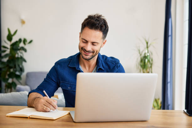 istock Man at home writing in notebook using laptop sitting at table in living room 2194654545