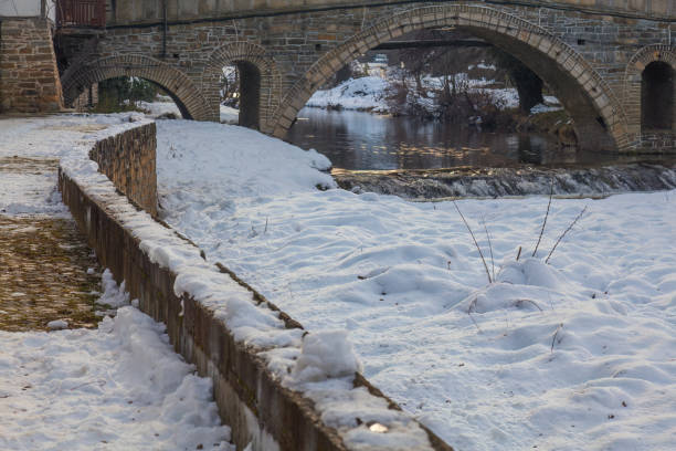 Old stone bridge over a river with snow on the ground, Tryavna The bridge is old and has a stone wall on the side