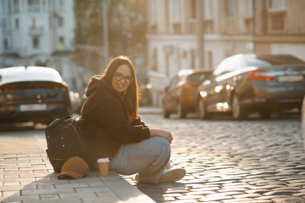 Model in brown coat, jeans and black boots sits in autumn city park on bench. against the background of old architecture. Vintage camera in hands, looking at camera