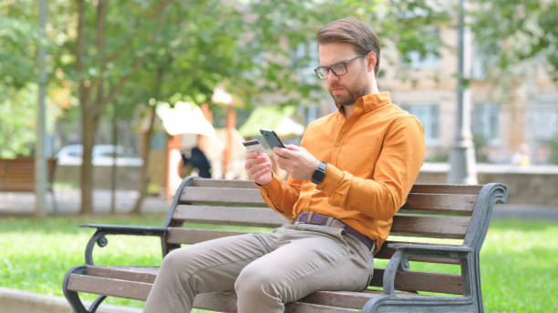 istock Young Man Doing Online Banking with Smartphone while Sitting on Bench 2194680894