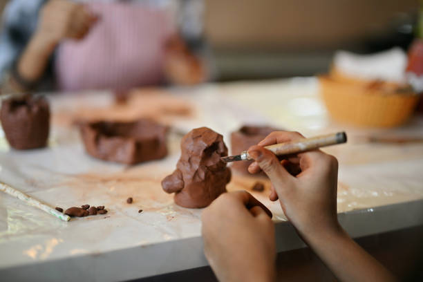 A cropped, detailed view of hands expertly molding terra-cotta clay using sculpting tools. The fingers, coated with earthy clay, move with precision and care, highlighting the craftsmanship and artistry involved. This close-up captures the tactile beauty and skill of the terracotta crafting process.