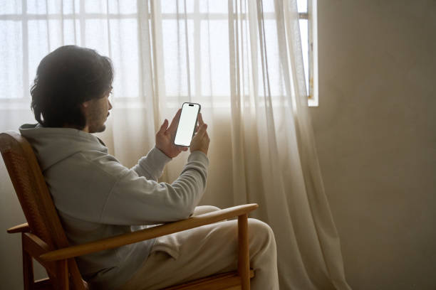 istock Young man relaxing on rocking chair using smartphone with blank screen by window 2194688671