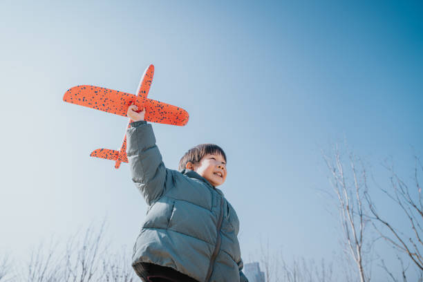 A Happy lovely 5-years-old  little Asian boy having fun outdoors, playing with airplane toy and smiling joyfully in park on a lovely sunny day against beautiful blue sky.