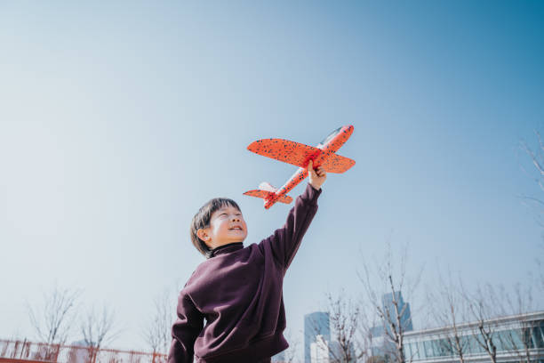 A Happy lovely 5-years-old  little Asian boy having fun outdoors, playing with airplane toy and smiling joyfully in park on a lovely sunny day against beautiful blue sky.