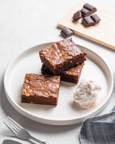 istock Chocolate brownie with nuts cut into pieces on a plate with whipped cream on a light background 2194691507