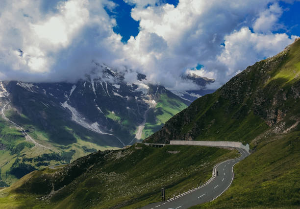 A winding mountain road cuts through lush green hills under a dramatic sky with clouds partially covering snow-capped peaks in the distance.