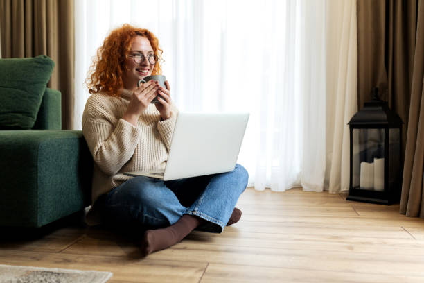 istock Young woman sitting on the floor in her living room while using laptop computer and drinking coffee 2194724747
