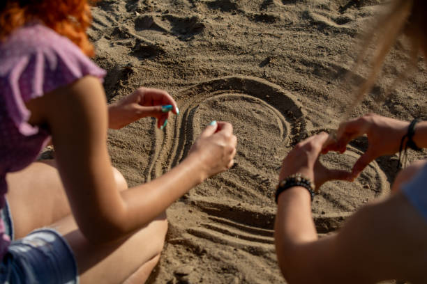 Two friends joyfully drawing patterns in the warm sand at a sunny beach, showcasing their creativity