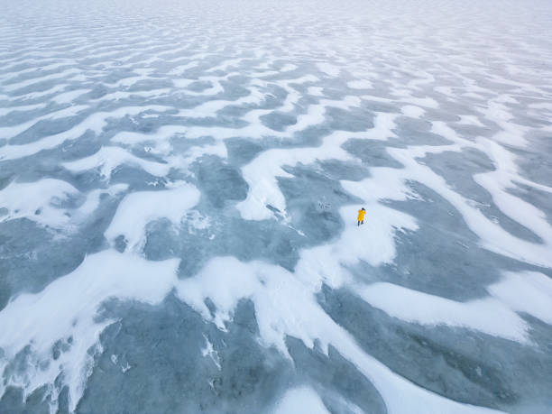 Aerial view of a scientist wearing a yellow coat walking on a frozen lake covered with snow and ice, conducting research in a polar environment