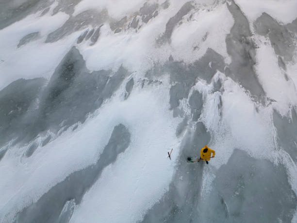 Aerial view of a scientist in yellow parka using specialized tools to extract ice core samples from a frozen lake surface, contributing to climate change research