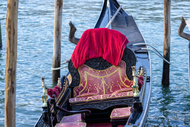 A beautifully decorated Venetian gondola floating on the calm waters, showcasing its intricate golden details and luxurious red velvet seating. The traditional wooden boat, moored between wooden poles, captures the essence of Venice’s romantic and historical charm. A perfect symbol of Italy’s rich cultural heritage and timeless beauty.