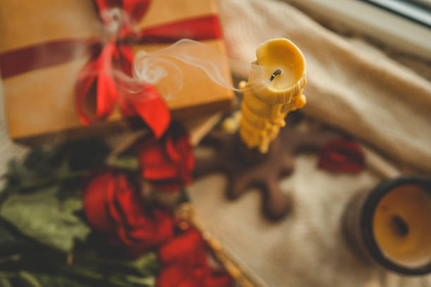 Twisted extinguished candle with soft swirling smoke in a cozy setting, accompanied by a kraft gift box tied with a red ribbon, red roses, and blurred decor creating a warm, romantic ambiance.