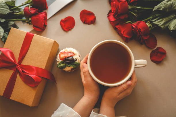 istock Child's hands holding a warm cup of tea surrounded by a gift box, red roses, and a cupcake on a beige surface. A cozy and heartfelt composition perfect for special occasions or celebrations 2194750708