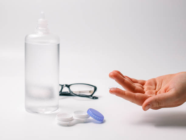 istock Hand holding a transparent contact lens next to lens case, solution bottle, and glasses on a white background. Concept of eye care, vision correction, and optical hygiene 2194751663