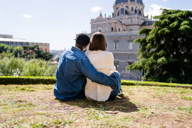 Multiethnic couple sitting on grass embracing and admiring a great cathedral in a city during sunny day