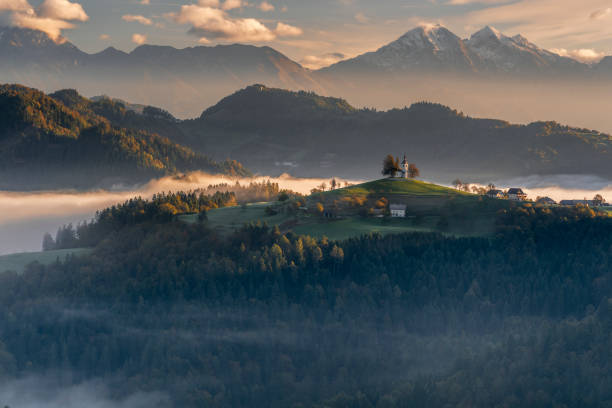A photo of the Church of St. Primus and Felician in Jamnik, surrounded by a sea of mist against the backdrop of Slovenia’s majestic mountains. The warm hues of the sunset/​sunrise sky lend a mesmerizing ambiance, ideal for travel, nature, and landscape-themed projects.