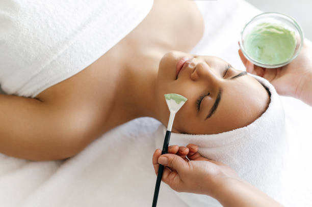 Facial treatments, wrinkle prevention, facial cleansing. Top view of beauty procedure, therapist applying green face mask on the face of a beautiful brazilian or hispanic young woman stock photo