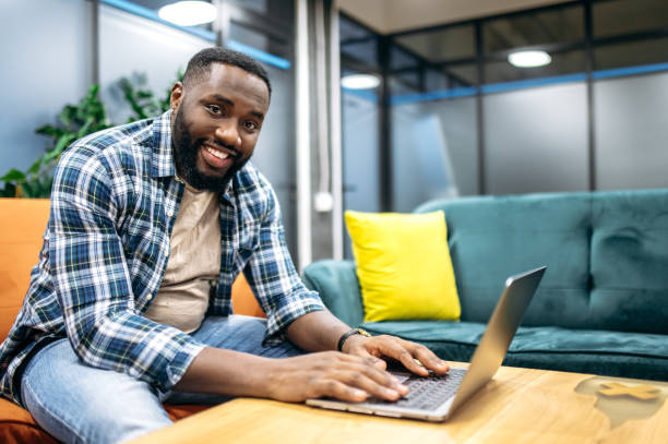 istock Joyful stylish freelancer sitting on the sofa, using laptop. Successful happy african american programmer looking at the camera, smiling, working on new project or app 2194794418