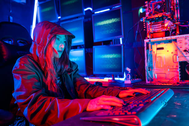 istock Woman hacker. Asian woman wearing hood is typing keyboard for coding hacking programming system. room is dimly lit. possibly typing or browsing internet, Hacker in neon light 2194803715