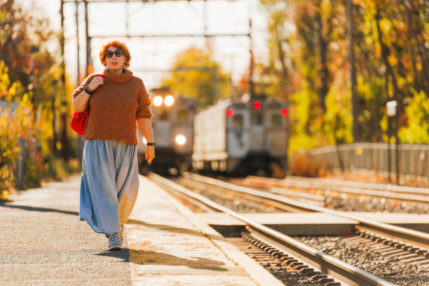 A 46-year-old woman with red curly hair stands poised on a transit platform as golden sunlight bathes the scene. Her brown turtleneck sweater and flowing denim maxi skirt create an elegant silhouette against the autumn backdrop. Sunglasses shield her eyes while a red handbag complements her style. Autumn trees frame the scene as approaching transit signals merge with sunset hues. Long shadows stretch across concrete while gleaming rails catch late day light.
