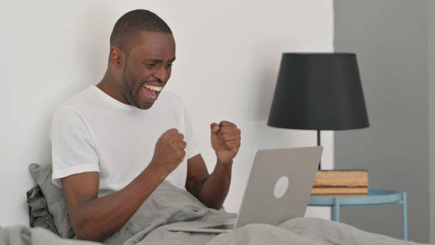istock African Man Celebrating Work on Laptop while Sitting in Bed 2194814057