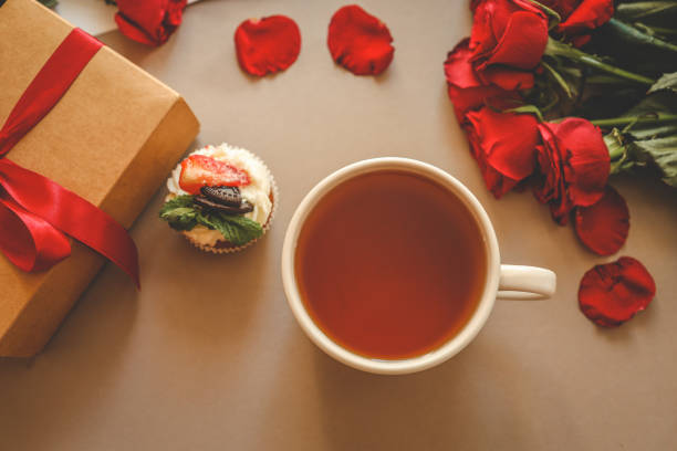 istock Flat-lay composition featuring a kraft gift box tied with a red ribbon, a cup of tea, a cupcake with fruit and mint topping, and vibrant red roses arranged on a beige background 2194815628